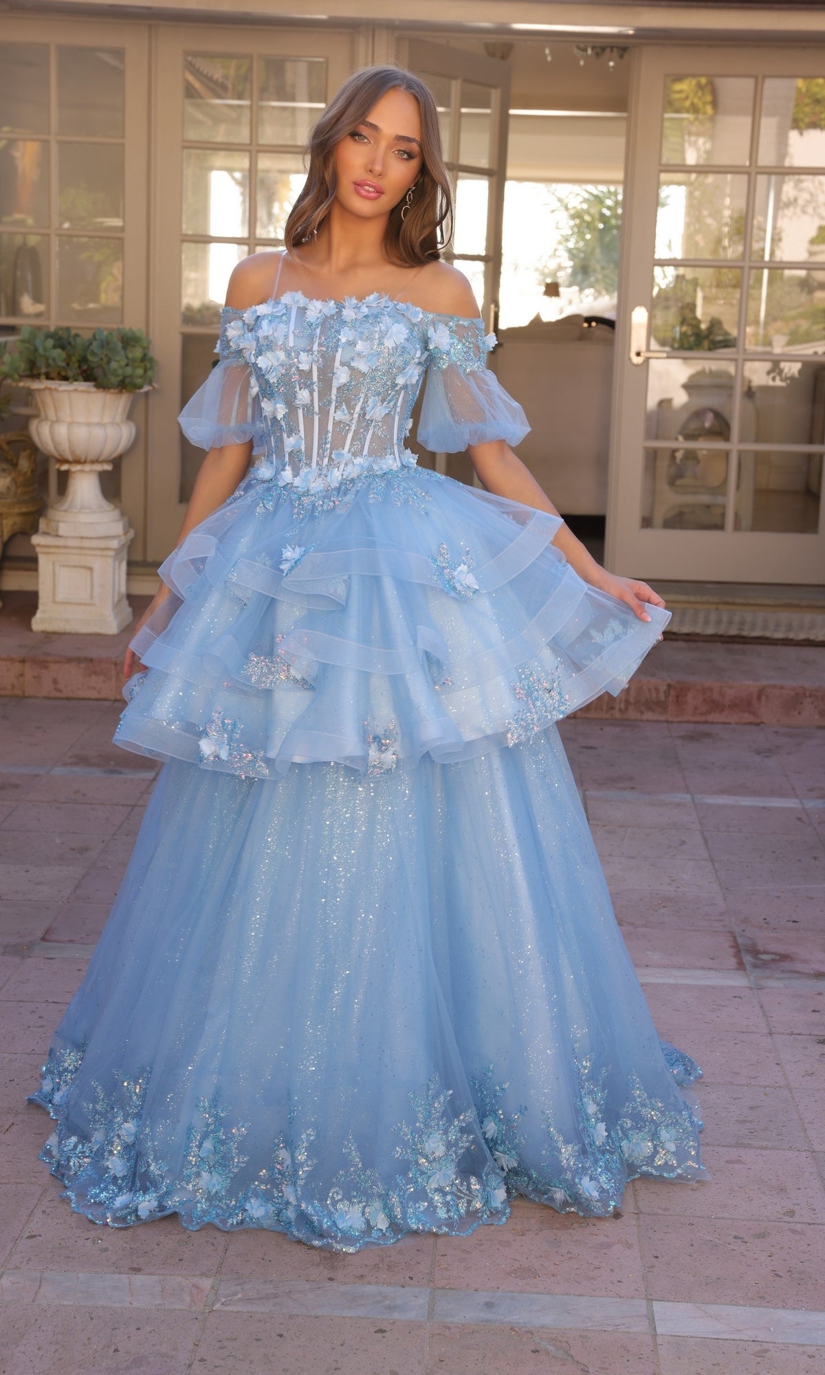 A woman stands outdoors in front of glass doors wearing the Nox Anabel Removable-Skirt Formal Ball Gown H1360, a light blue off-the-shoulder dress featuring floral details, a layered tulle skirt, and sheer overlay.